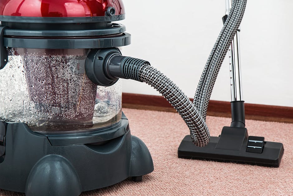 Close-up view of a vacuum cleaner with a transparent dust container filled with debris, connected to a flexible, patterned hose, positioned on a pinkish-beige carpet in a room with a white wall and wo